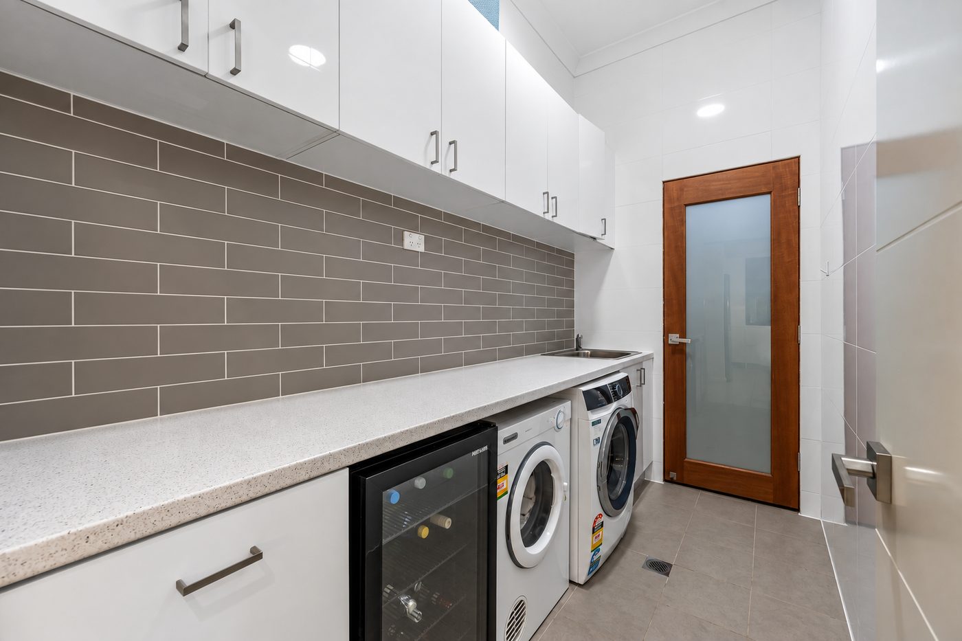 Moorebank laundry with a long bench, grey tile splashback, wine fridge and timber-framed frosted door